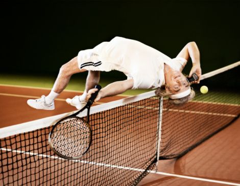 Ældre herre flyver på hovedet over tennisnettet, imens han taler i telefon og slår til bolden samtidigt.